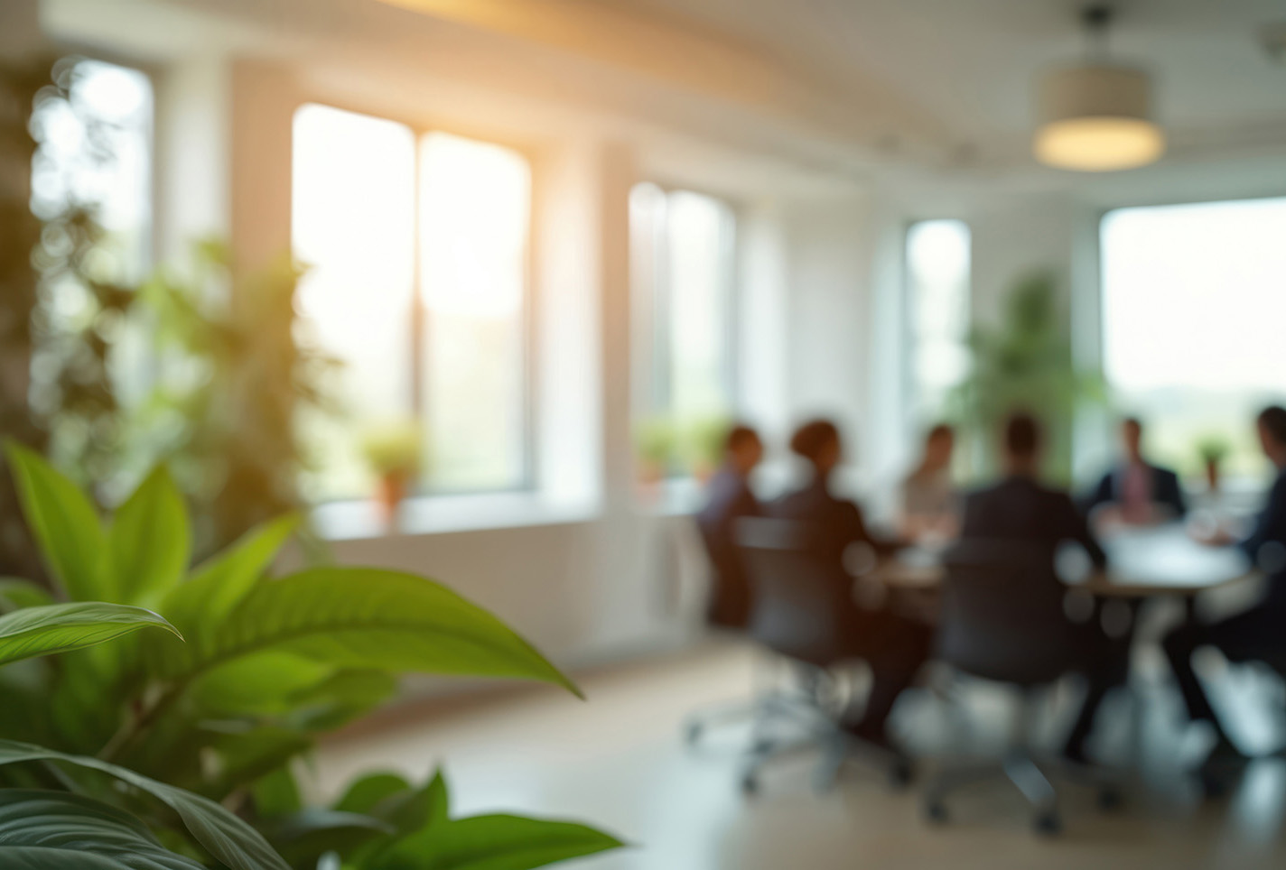 Business people meet around table in bright modern office. Green plants add life to meeting room setting. Natural light fills the space through large windows.